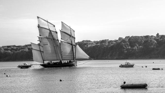 Black and white photo of a two-masted vintage sailing ship, sail number CAN 87, with crew on deck, on calm water near a wooded shore, with small motorboats and buoys around it.
